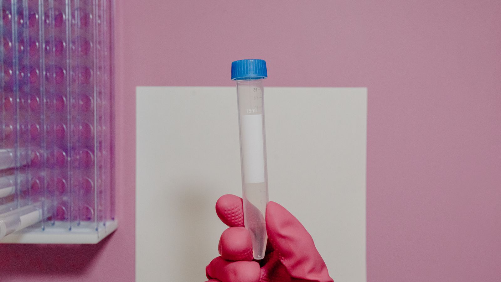 A gloved hand holding a test tube in a laboratory setting with a pink background.