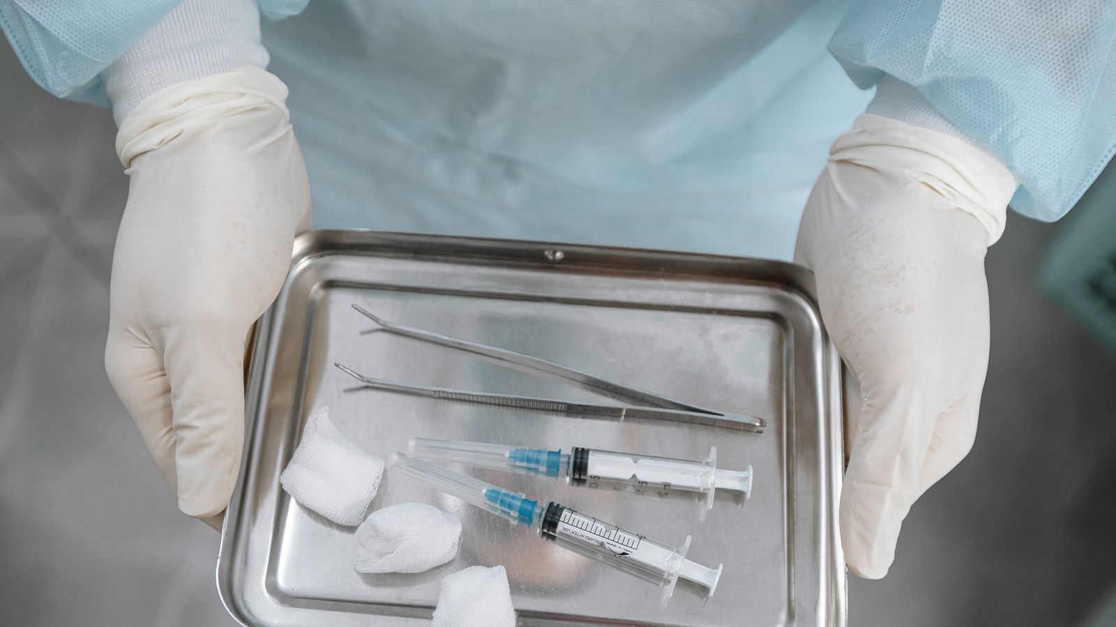 Close-up of a medical professional's gloved hands arranging syringes, tweezers, and gauze on a steel tray.