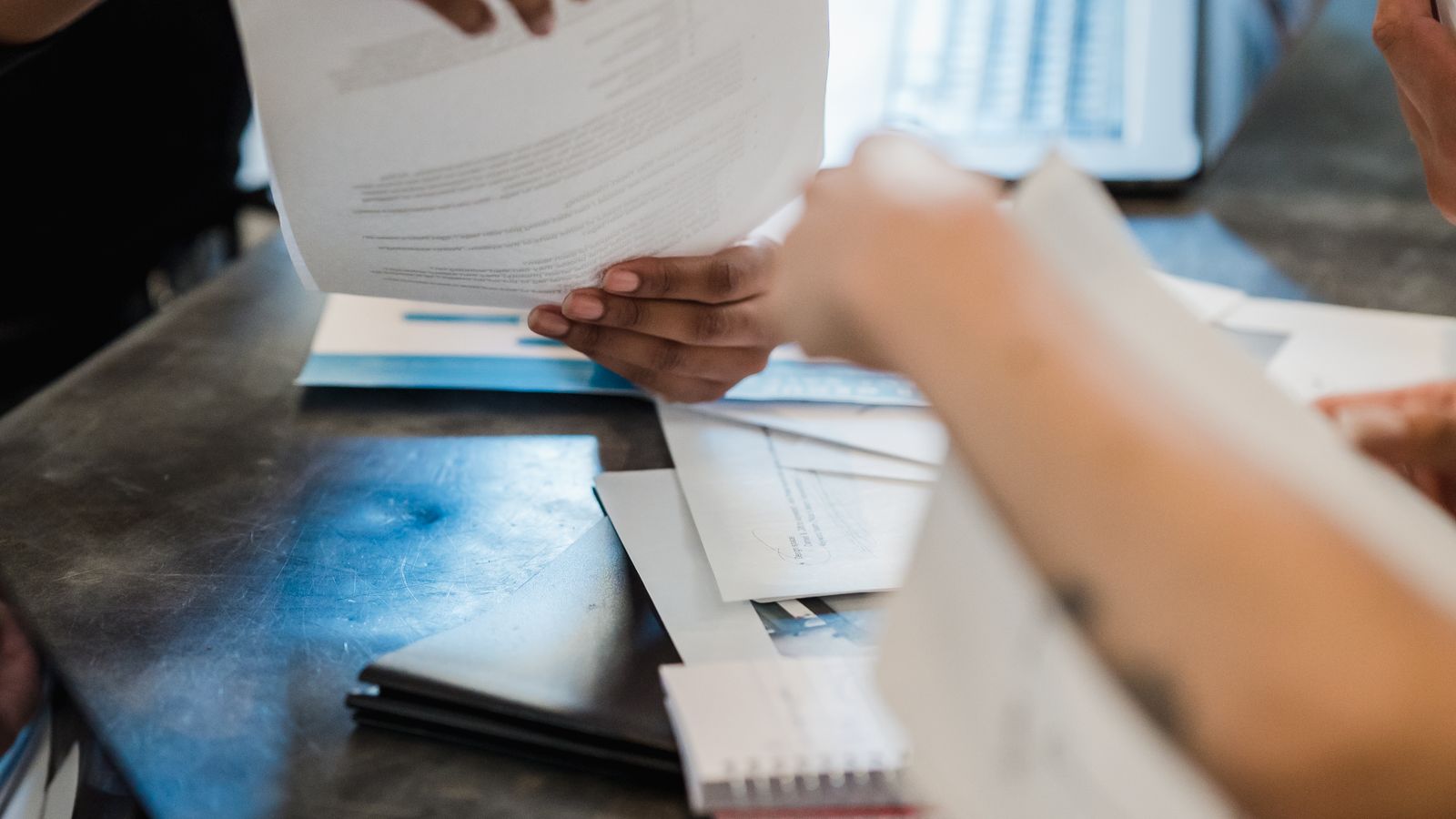 Two people exchanging paperwork across a table while a laptop sits open between them in an office setting.