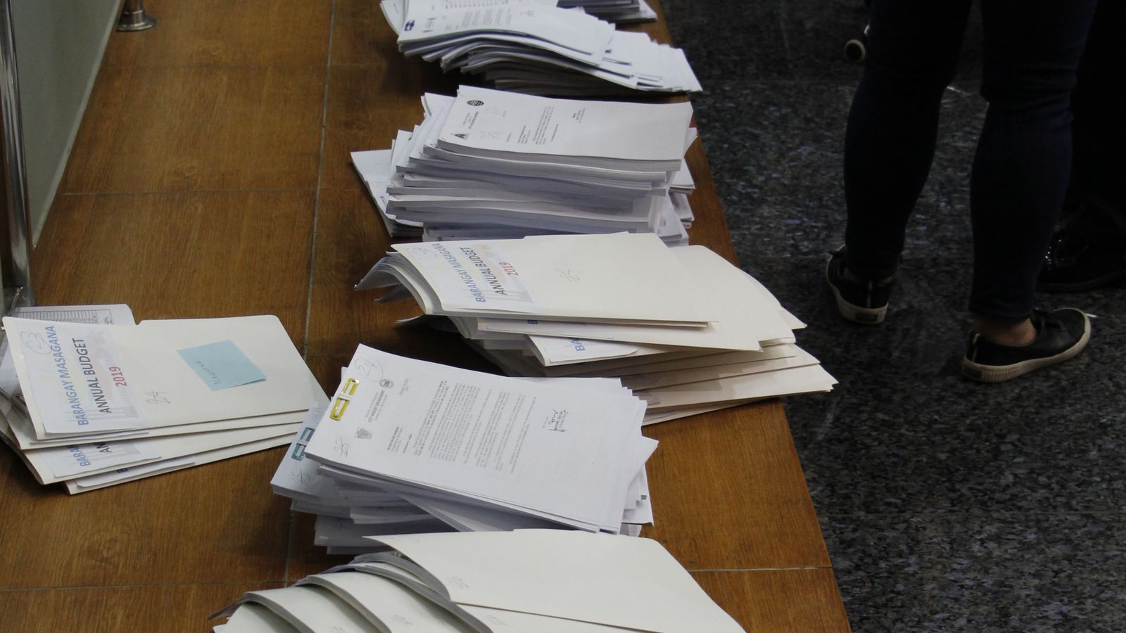 Stacks of paper documents stacked neatly on a wooden desk in an office setting.
