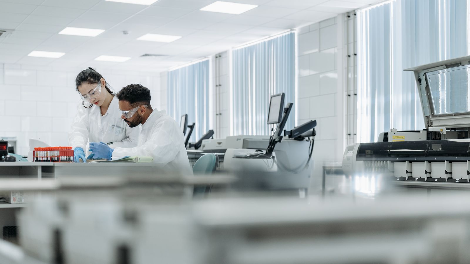 Two scientists in a laboratory wearing protective eyewear and gloves while examining research materials together.